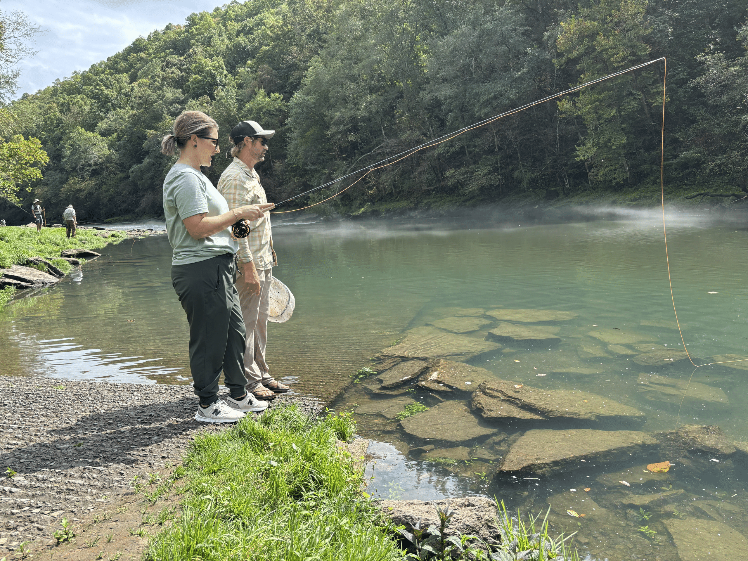 Woman standing with man at the bank of a river. She is learning how to fish and is holding a reel in her hand.